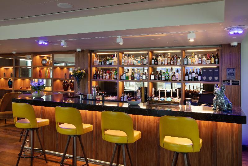 Tower Suites By Blue Orchid Лондон Екстериор снимка The photo shows a modern bar area. In the foreground, there are several yellow bar stools lined up in front of a sleek black countertop. Behind the bar, there is a well-stocked shelf displaying a variety of bottles, including spirits and liqueurs, il