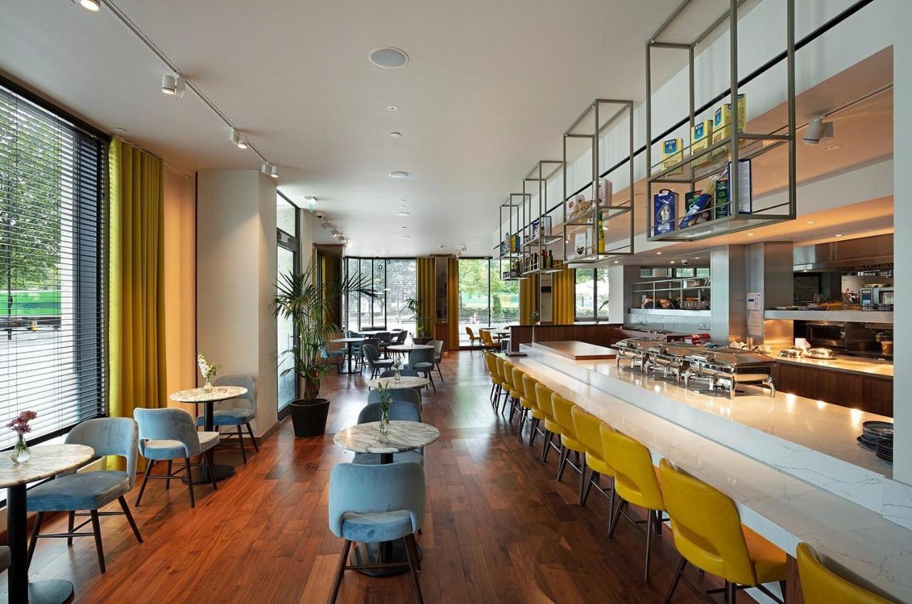 Tower Suites By Blue Orchid Лондон Екстериор снимка The photo shows a modern restaurant interior. It features a spacious layout with large windows allowing natural light to flood the space. The decor includes a combination of wooden flooring and colorful furniture—blue and yellow chairs arranged aroun