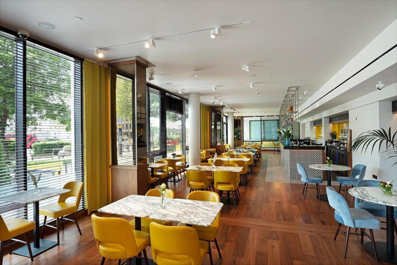 Tower Suites By Blue Orchid Лондон Екстериор снимка The photo depicts a modern dining area with a bright and airy atmosphere. There are several tables with marble tops, accompanied by yellow chairs, creating a vibrant contrast with the wood flooring. The space features large windows that allow ample n