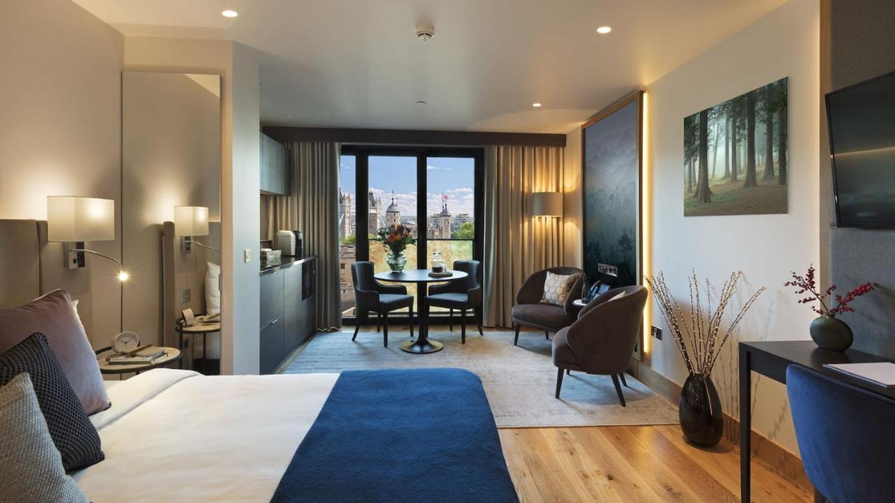 Tower Suites By Blue Orchid Лондон Екстериор снимка The photo shows a modern hotel room designed with a contemporary aesthetic. On one side, there is a large bed with a blue blanket, while the other side features a sitting area with two chairs and a small round dining table. Large windows let in natur