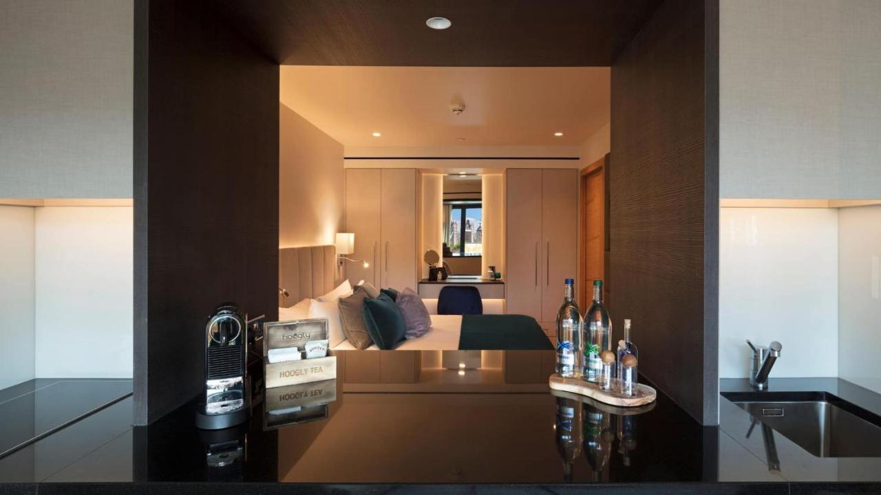 Tower Suites By Blue Orchid Лондон Екстериор снимка The image shows a modern hotel room with a stylish design. In the foreground, there is a dark countertop with several bottles displayed, likely including spirits and water. Just beyond the countertop, you can see the bedroom area, which has light-col