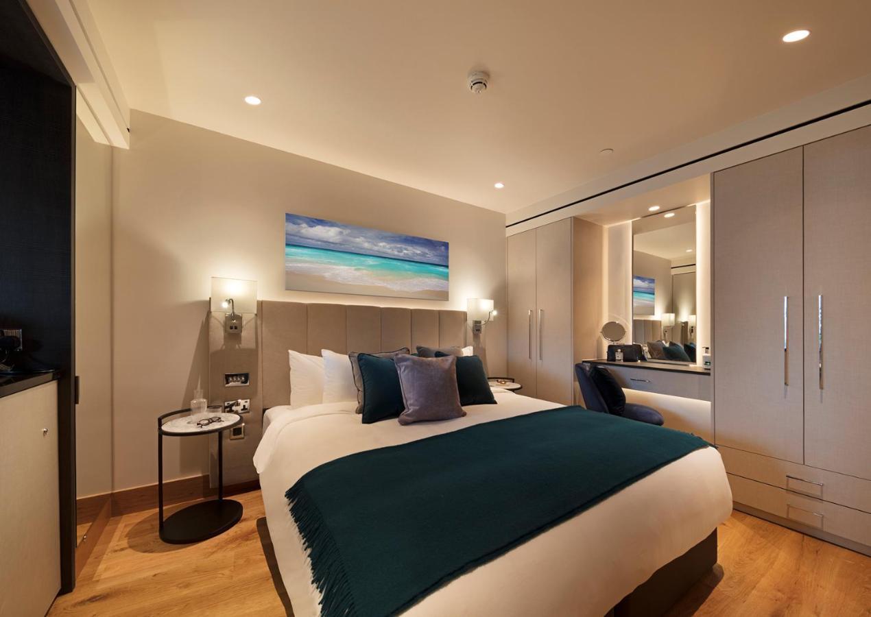 Tower Suites By Blue Orchid Лондон Екстериор снимка The photo shows a modern hotel room featuring a double bed with a white duvet and a dark teal throw blanket. There are decorative pillows in shades of blue and purple on the bed. To one side, there's a round bedside table with a small lamp. A large m