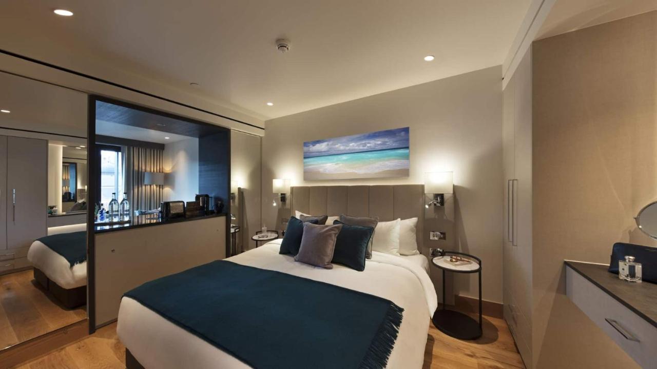 Tower Suites By Blue Orchid Лондон Екстериор снимка The photo shows a modern hotel bedroom featuring a large bed with a light-colored headboard and a deep blue throw at the foot. The bed is adorned with multiple decorative pillows. On the wall above the bed, there is a colorful painting depicting a be