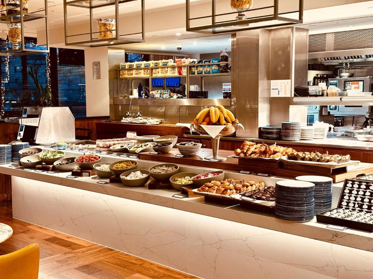 Tower Suites By Blue Orchid Лондон Екстериор снимка The photo depicts a buffet-style setup, likely in a restaurant or a hotel dining area. In the foreground, there are multiple bowls and platters filled with a variety of food items, including salads, fruits, pastries, and other dishes. There is a long
