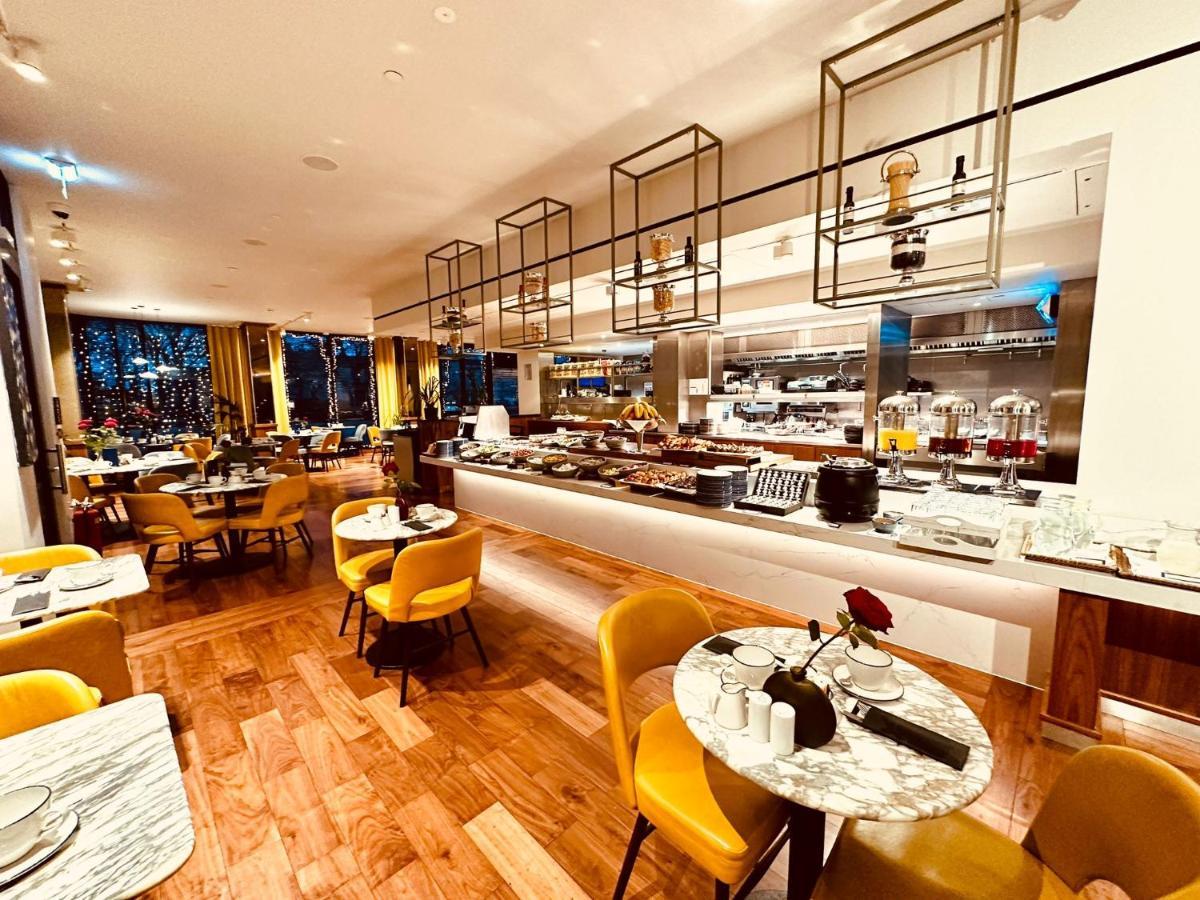 Tower Suites By Blue Orchid Лондон Екстериор снимка The photo depicts a modern restaurant interior with a spacious layout. There are several round tables, each adorned with marble tabletops and stylish yellow chairs. A buffet area is visible in the background, featuring various food setups and kitchen